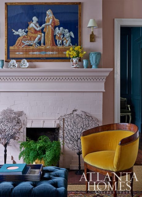 Living room with a pink brick fireplace, a yellow velvet armchair, and a blue tufted ottoman. A tapestry hangs above the mantel.