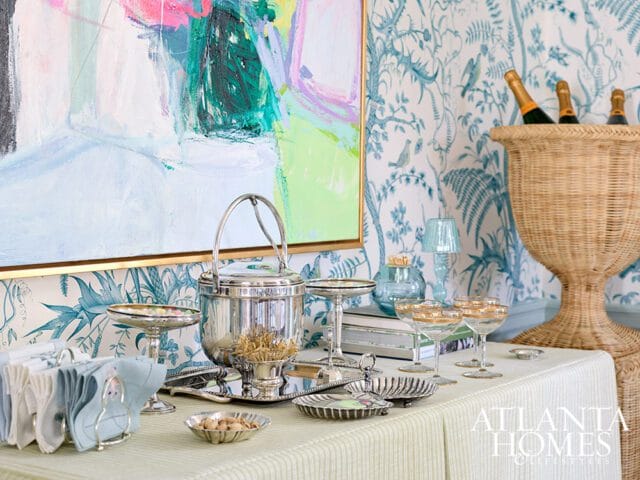 A bar setup with a silver ice bucket, cocktail glasses, and a woven champagne bucket against a blue botanical wallpaper.