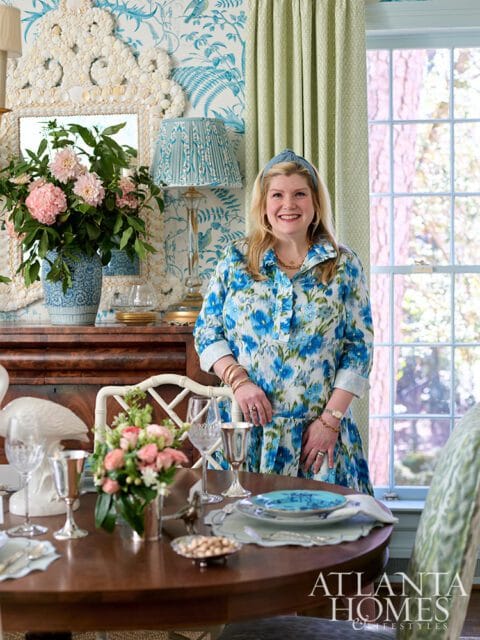 Woman in a blue floral dress standing in a dining room with blue and white bird-themed wallpaper and green curtains.