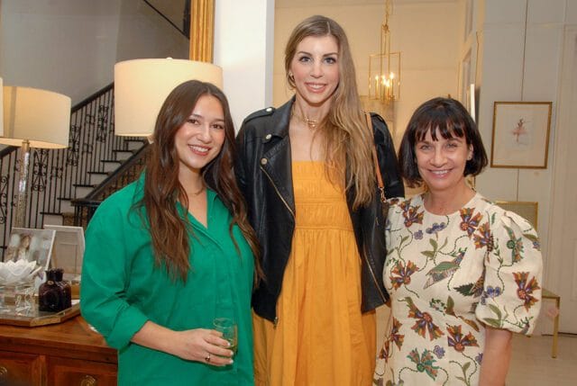 Three women smiling indoors. The woman on the left wears a green shirt and holds a glass. The woman in the middle wears an orange dress and black leather jacket. The woman on the right wears a cream-colored dress with a floral pattern.