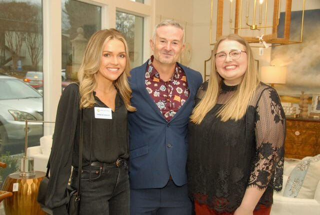 Three people posing for a photo indoors. The person on the left wears a black shirt and jeans, the person in the middle wears a blue suit jacket over a patterned shirt, and the person on the right wears a black lace top.