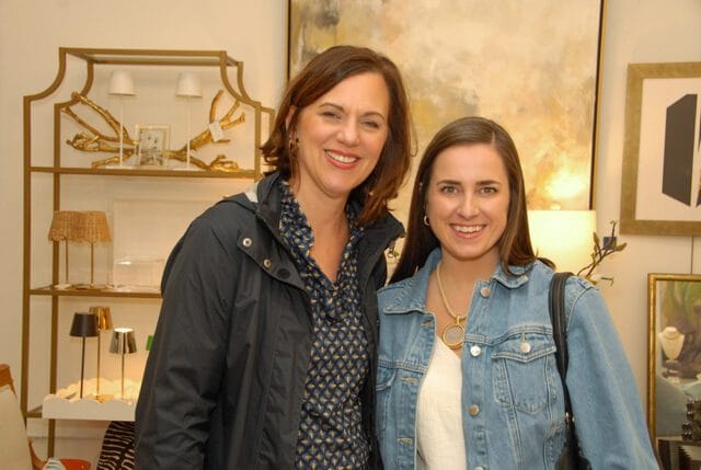 Two women smiling at the camera in a room with artwork and decorative shelving. The woman on the left wears a dark jacket over a patterned blouse. The woman on the right wears a denim jacket over a white top.