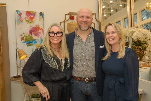 Three people smiling for a photo in a room with artwork and decorative objects. The woman on the left wears glasses and a black ruffled blouse. The man in the middle wears a patterned shirt and navy blazer. The woman on the right wears a navy dress.