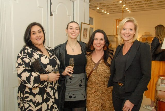 Four women posing for a photo at an event. The woman on the far left wears a black and white floral dress. The second woman from the left wears a black top and skirt with a leather jacket. The third woman wears a patterned brown dress. The woman on the far right wears a black blazer and pants.