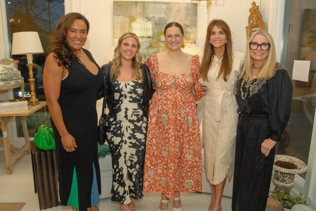 Five women stand together in a room with artwork on the wall and decorative items in the foreground.