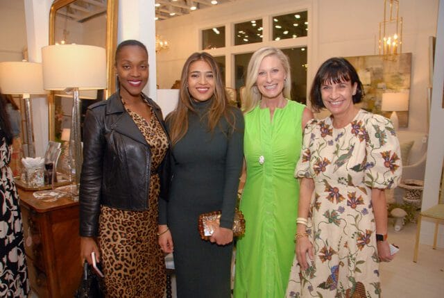 Four women smiling at the camera in a well-lit room with decorative lamps and furniture.