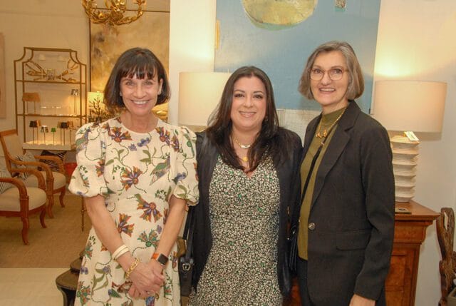 Three women smiling at the camera in a room with decorative furniture and artwork. The woman on the left wears a floral dress, the middle woman wears a patterned dress and black jacket, and the woman on the right wears a dark blazer over a green turtleneck.