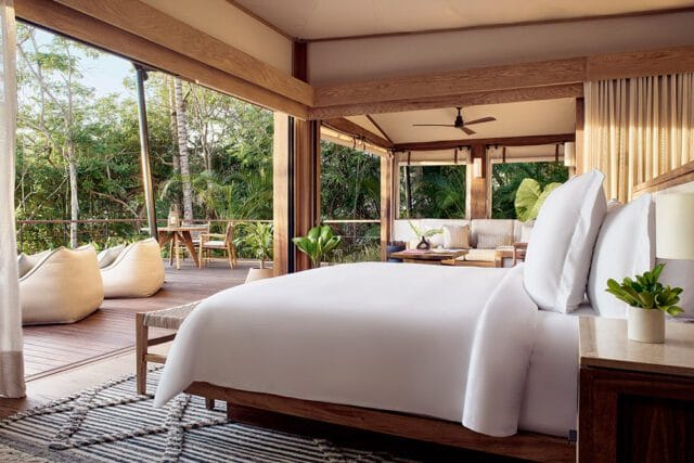 Bedroom with white bedding, wooden beams, and large glass doors opening to a lush green outdoor space with seating.