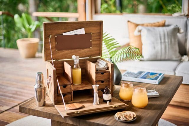 Wooden bar chest open on a table, containing bottles of liquid, cocktail shaker, jigger, and glasses with orange drinks. A book and dried fruit are on the table.