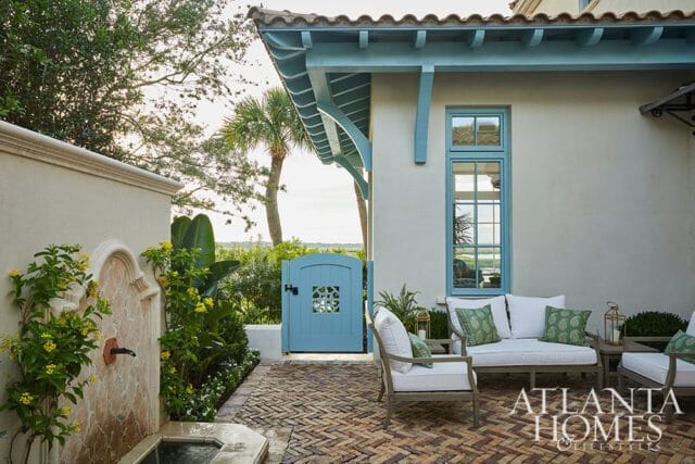 Courtyard with a stone fountain, herringbone brick patio, light-colored stucco walls, and a light blue arched gate. Outdoor seating with white cushions and green patterned pillows is visible.
