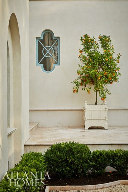Cream stucco wall with a light blue quatrefoil window and a potted citrus tree with orange fruits.