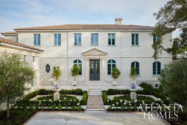 Mediterranean-style villa with white stucco facade, terracotta tile roof, and light blue-trimmed windows, featuring a formal garden and stone pathway leading to the entrance.