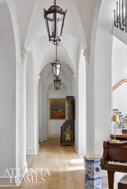 White arched hallway with hanging lanterns, a dark ornate cabinet, a landscape painting, and a piano