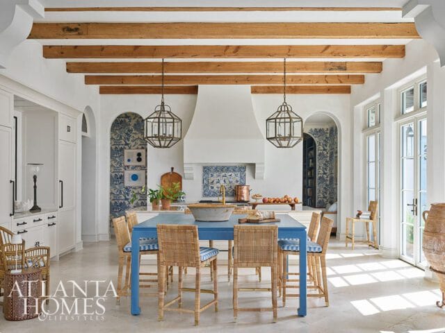 Kitchen with blue island, rattan bar stools, white cabinetry, exposed wood beams, and blue and white patterned wallpaper.