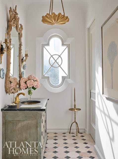 Powder room with a distressed wood vanity, black granite countertop, brass faucet, and a decorative mirror. Features a unique arched window, gold fan-shaped pendant light, and a patterned tile floor.