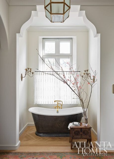 Freestanding dark metal bathtub with brass fixtures in a white bathroom with a window, ornate sconces, and a decorative chest side table.