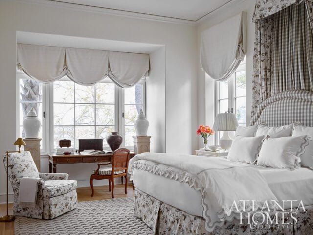 Bedroom with a large window featuring draped valances, a desk with a chair, a floral armchair, and a bed with a gingham headboard and floral skirt.