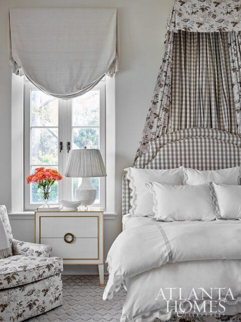 Bedroom with a white and gray gingham canopy bed, white bedding with scalloped trim, a floral armchair, a white nightstand with gold accents, and a window with a draped valance.