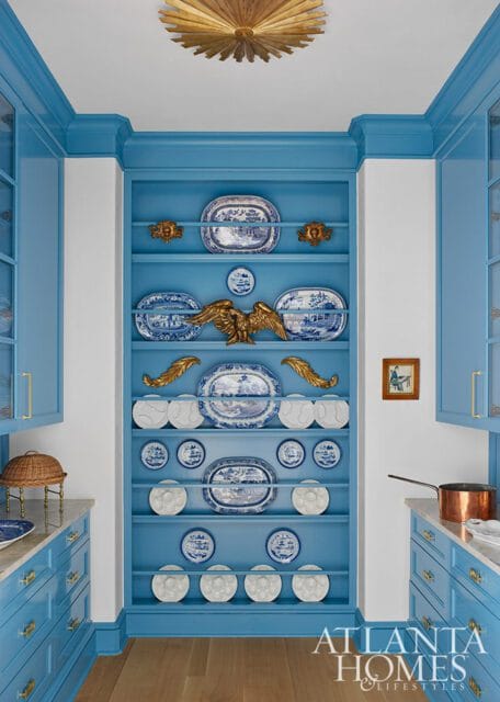 Blue kitchen with open shelving displaying blue and white plates, gold eagle sculpture, and brass hardware. A copper pot sits on the counter.