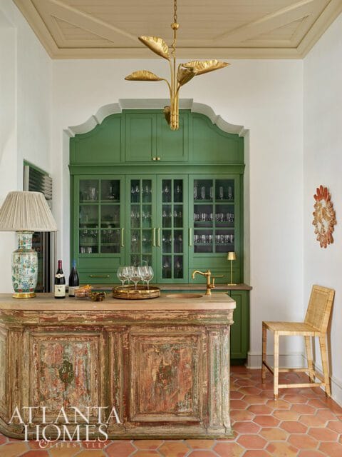 Bar area with green built-in cabinetry, distressed wood bar, brass faucet, rattan stool, and terracotta tile floor.