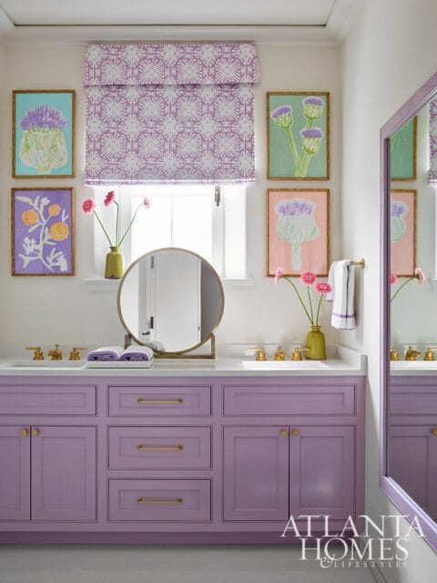 Purple vanity with brass hardware, white countertops, and a round mirror. Artwork of thistles and oranges adorn the wall above the vanity.