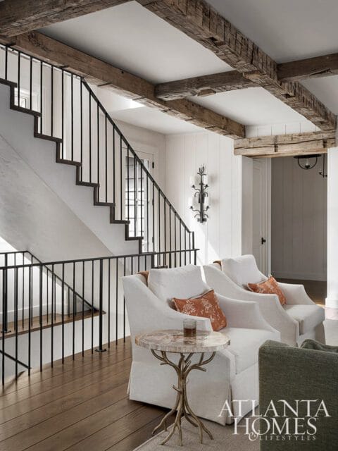 Living room with white slipcovered armchairs, rustic wooden beams on the ceiling, a black metal staircase railing, and a small side table with a petrified wood top.