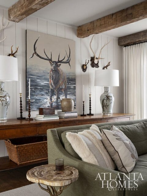 Living room with rustic wooden beams, a large painting of an elk, mounted antlers, a wooden console table, and a green sofa with decorative pillows.