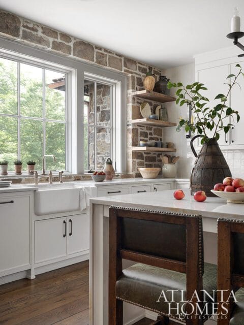 Rustic stone accent wall in a white kitchen with a farmhouse sink, marble island, and wooden bar stools.