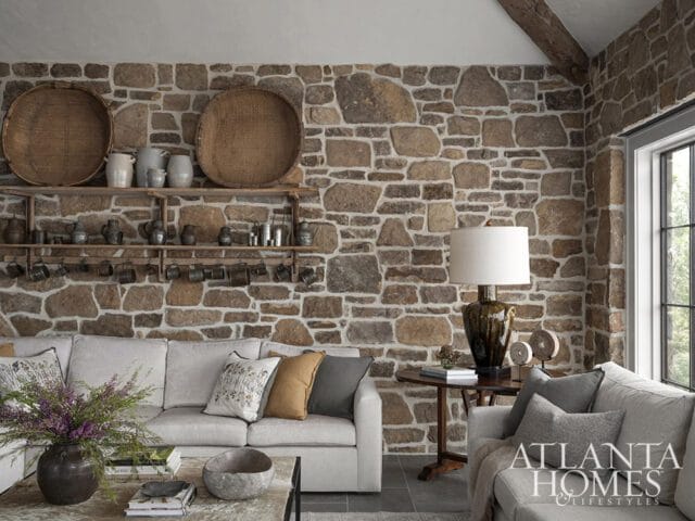Living room with a stone accent wall, light gray sectional sofa, wooden shelves with pottery and baskets, and a round side table with a lamp.