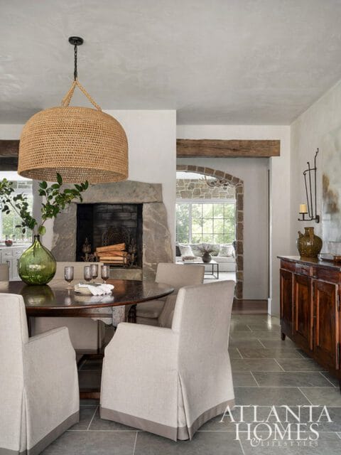 Dining room with a round wooden table, upholstered chairs, stone fireplace, and a large woven pendant light.