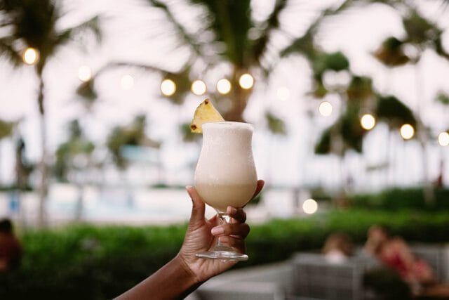 Hand holding a frosted glass filled with a creamy, light-colored cocktail garnished with a pineapple wedge. String lights and palm trees are blurred in the background.