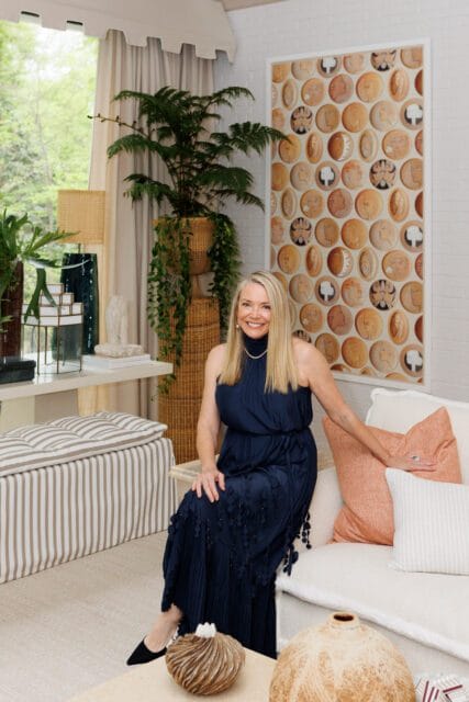 Woman in navy dress sitting on a white sofa in a living room with a large framed artwork of circular portraits on the wall and a striped ottoman.