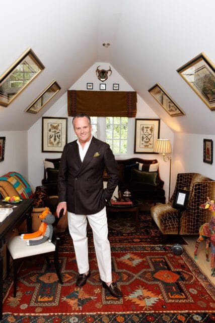 Man in dark suit and white pants standing in a room with slanted ceilings, oriental rugs, and eclectic decor including taxidermy and framed insect prints.