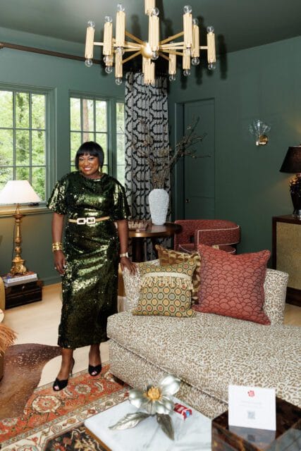 Woman in a green sequined dress stands in a room with dark green walls, a gold chandelier, patterned curtains, and a leopard-print chaise lounge.