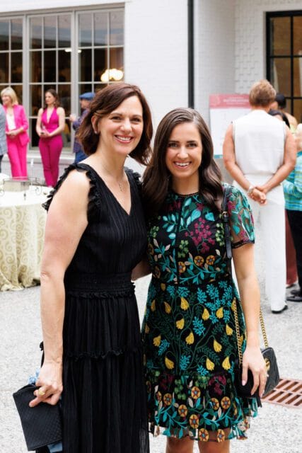 Two women smiling at the camera, one in a black dress and the other in a black dress with colorful floral embroidery.