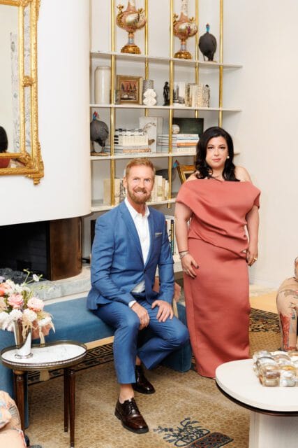 Man in blue suit and woman in coral dress pose in a living room with a fireplace, gold-trimmed shelves filled with decorative objects, and patterned rugs.