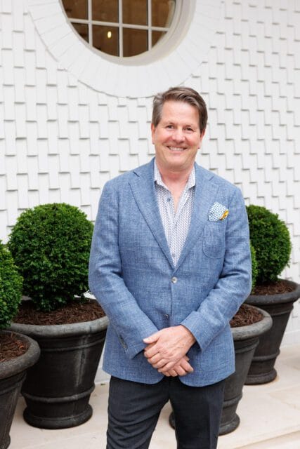 Man in blue blazer and patterned shirt smiling in front of white textured wall with round window and potted boxwood plants.