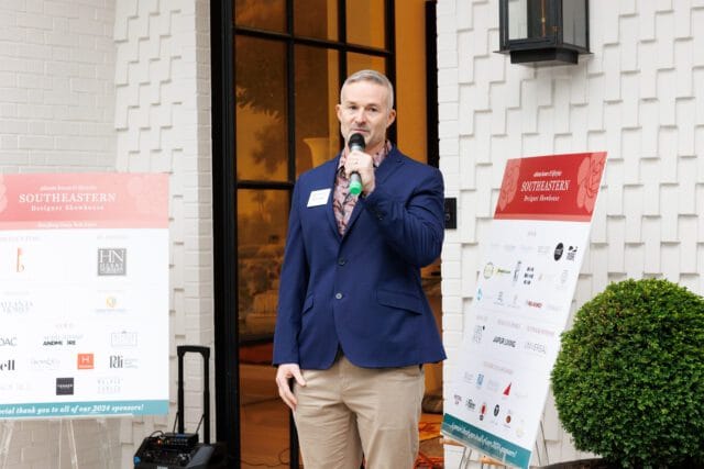 Man in navy blazer speaking at podium next to signs for Southeastern Designer Showhouse benefiting Camp Twin Lakes, listing sponsors.
