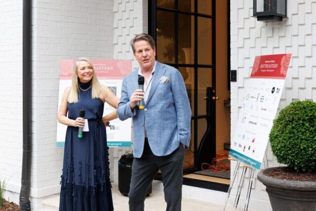 A woman in a navy dress and a man in a blue blazer speak into microphones at the Southeastern Designer Showhouse opening, with sponsor signs behind them.