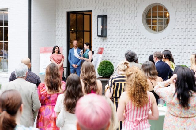People gathered outside a white house with textured siding, a circular window, and a black lantern. Three people stand near a podium speaking to the crowd.