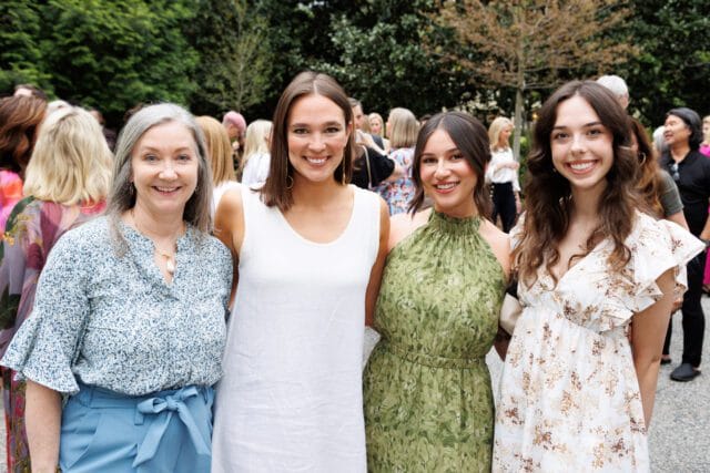 Four women smiling at the camera, with trees and a crowd in the background. The woman on the far left wears a blue floral blouse and blue pants. The next woman wears a white sleeveless dress. The third woman wears a green halter dress. The woman on the far right wears a white floral dress.