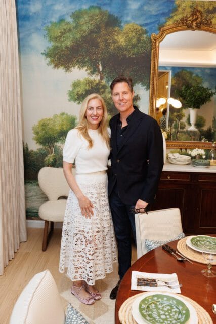 A man and woman pose in a dining room with a scenic mural wallpaper and a large ornate mirror.