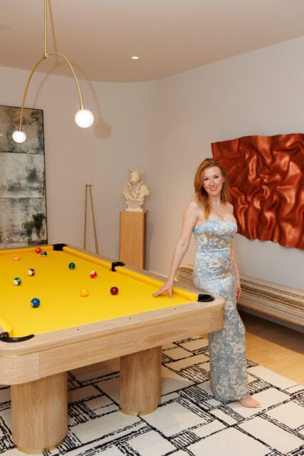 Woman in a blue floral dress leans on a light wood pool table with a bright yellow felt. Modern art hangs on the wall.