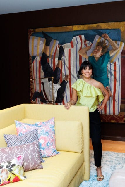 Woman in a lime green top and black pants standing next to a yellow sofa with patterned pillows. Behind her is a large painting of figures on a striped bed.