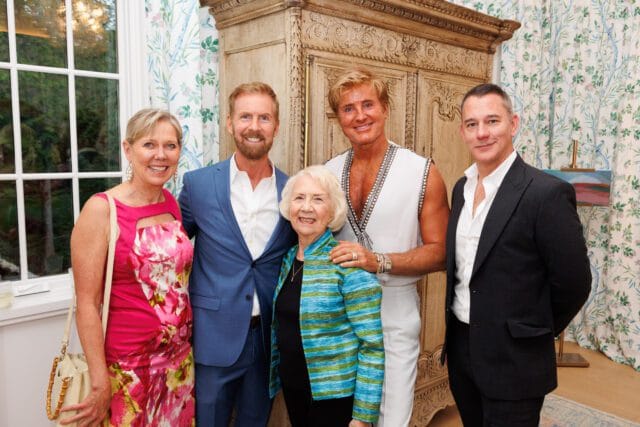 Four people pose for a photo in a room with floral wallpaper and a large wooden armoire. A woman in a pink dress, a man in a blue suit, an older woman in a striped jacket, and a man in a white sleeveless top and dark suit.
