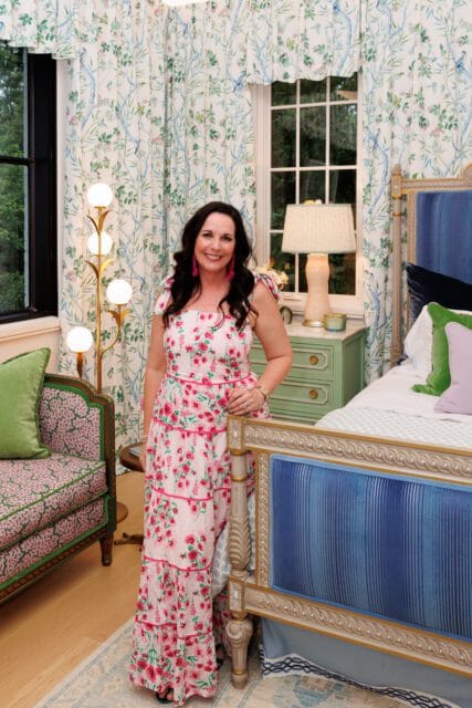 Woman in floral dress in a bedroom with blue velvet bed, green accent chair, and botanical print curtains.