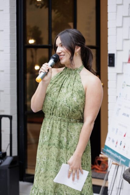 Woman in a green floral dress holding a microphone and papers, speaking at an event.