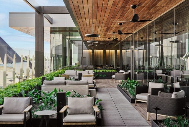 Modern lounge area with plush gray seating, wood-paneled ceiling with black ceiling fans, and floor-to-ceiling glass windows overlooking an outdoor terrace.