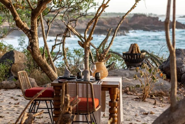 Table set for two on a sandy beach with ocean views, featuring wicker chairs with orange cushions, wooden table, glassware, and a fire pit in the background.