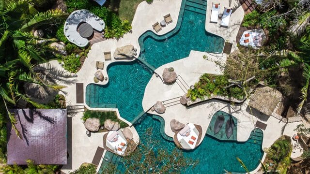 Aerial view of a resort with a winding turquoise pool, lounge areas, thatched roofs, and lush tropical foliage.